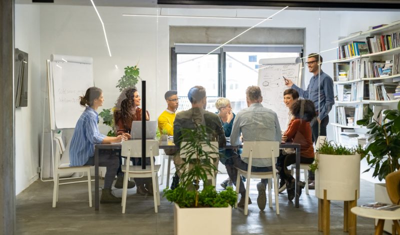 Confident mature businessman giving a presentation to his crative team in office. Business brief with annual goals with casual employees. Happy leadership man training young businessmen and smiling businesswomen sitting at conference table.